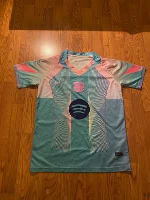 Barça x Nike pastellfärgad matchtröja - Säljer en FC Barcelona matchtröja från Nike i pastellfärger med mönster i turkos, rosa och ljusblått. Tröjan har klubbmärke och sponsorlogga på bröstet samt korta ärmar och sportig passform. Perfekt för dig som älskar fotboll och snygg design.