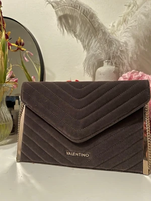 Mörkbrun kuvertväska från Valentino - Snygg mörkbrun kuvertväska från Valentino i mjuk sammet med guldfärgade metalldetaljer och kedjerem. Väskan har ett elegant, vadderat mönster i chevron-stil och logga framtill. Perfekt accessoar för att lyfta din outfit.