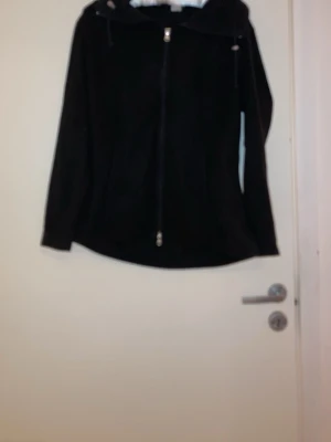 Svart fleecejacka med dragkedja - Mysig svart fleecejacka med huva och hel dragkedja framtill. Jackan har en enkel och stilren design med långa ärmar och lätt utsvängd passform. Perfekt för kyliga dagar och enkel att matcha med olika outfits.