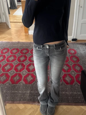Grå bootcut jeans med vita sömmar - Säljer ett par grå bootcut jeans med markerade vita kontrastsömmar och dekorativa fickor både fram och bak. Jeansen har låg midja och klassisk femficksdesign. Perfekta för en avslappnad och trendig look.