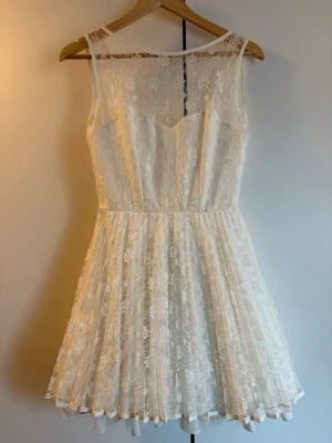 Vit spetsklänning med v-ringad rygg - Söt vit kortklänning i spets med ärmlös design och v-ringad rygg som knyts med ett band. Klänningen har ett tunt innerfoder och ett genombrutet spetslager över hela. Perfekt för sommarens alla tillfällen! Från Jones and Jones. Stl 10