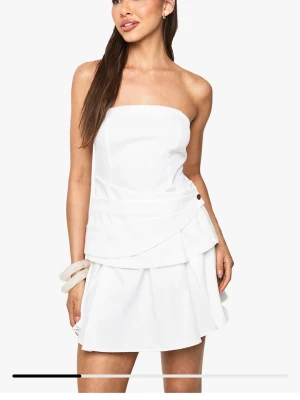 student klänning - Axelbandslös vit klänning med kort längd och volangdetaljer. Klänningen har en stilren och modern design. Endast testat 1 gång