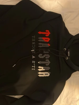 Svart hoodie från Trapstar - svart hoodie från Trapstar med huva och snören. Tryck i rött, vitt och grått på bröstet med texten 'Triumstar It's a Secret'. Mjuk insida och klassisk passform, perfekt för dig som gillar streetwear och vill sticka ut. Mindre i storlek så passar även M bra.