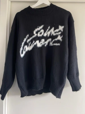 Svart stickad Saint Laurent tröja - Svart stickad tröja från Saint Laurent med vit, grafisk logga framtill. Tröjan har rund halsringning, långa ärmar och ribbade muddar vid ärmslut och nederkant. Materialet ser mjukt och lite luddigt ut, perfekt för kyligare dagar.