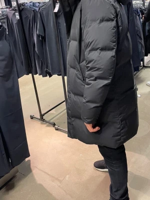 Svart lång dunjacka med huva - Säljer en svart lång dunjacka med huva och dragkedja framtill. Jackan har en stilren design med quiltade paneler och två sidofickor. Perfekt för kalla vinterdagar och håller dig varm och bekväm. Passar dig som gillar en enkel och modern look.