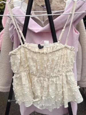 Krämvit volangig crop top - Säljer en söt krämvit crop top med tunna axelband och volangdetaljer. Toppen har en strukturerad yta med små tofsar och rynkade partier som ger en romantisk känsla. Perfekt att styla med jeans eller kjol för en trendig look.
