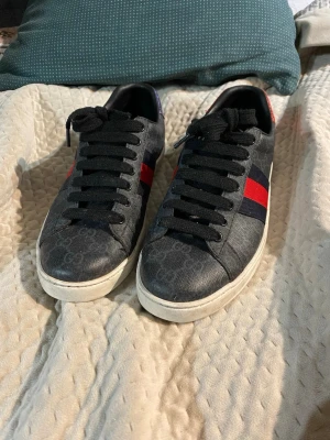 Gucci svarta sneakers med röd/blå detalj - Svarta sneakers från Gucci med diskret GG-mönster över hela skon och klassiska röda och blå ränder på sidorna. Skorna har svarta skosnören och vit sula. Perfekta för dig som vill ha en lyxig och stilren look.