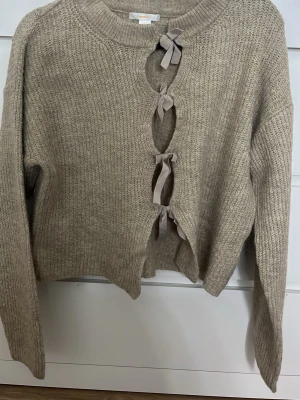 Beige stickad tröja med rosettknäppning - Trendig beige stickad tröja med unika öppningar framtill som stängs med tre dekorativa rosetter. Tröjan har rund halsringning, långa ärmar och ribbstickade muddar. Perfekt för dig som vill ha en mysig men ändå stilren look.