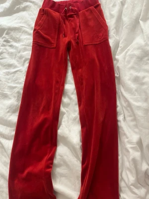 Röda velourbyxor Juicy Couture - Säljer ett par klassiska röda mjukisbyxor från Juicy Couture i mjuk velour. Byxorna har bred resår i midjan med dragsko, stora fickor fram och bakfickor med knappar. Perfekta för en chill och trendig look. Dom är lite lite hårda där nere efter användning men knappt något.