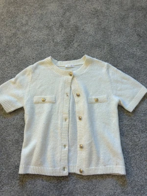 Vit kortärmad kofta Gina Tricot - Kortärmad kofta i mjukt vitt stickat material från Gina Tricot. Rund halsringning, dekorativa guldfärgade knappar framtill och två fejkfickor på bröstet. Ribbstickade kanter vid ärmslut, hals och nederkant. Perfekt för en clean och stilren look.