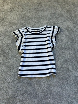 Randig topp med volangärm från Only Young - Snygg vit topp med svarta horisontella ränder från Only Young. Modellen har korta volangärmar och rund halsringning. Perfekt för en trendig och avslappnad stil. Materialet känns mjukt och bekvämt mot huden.