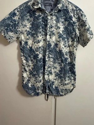 Blåvit mönstrad skjorta Vintage One - Kortärmad skjorta från Vintage One med blått och vitt tropiskt mönster. Skjortan har knappar framtill och klassisk krage. Perfekt för sommarens alla äventyr och ger en avslappnad vibe.