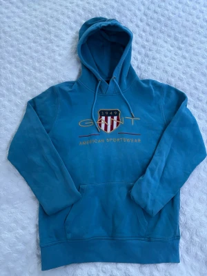Blå hoodie från GANT - Snygg blå hoodie från GANT med broderad logga och texten 'American Sportswear' på bröstet. Klassisk huva med dragsko och stor magficka framtill. Materialet är mjukt och skönt, perfekt för en avslappnad stil.