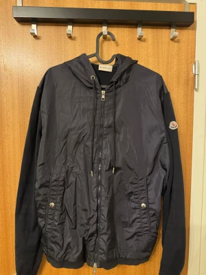 Moncler Cardigan - Snygg mörkblå vindjacka från Moncler med huva och dragkedja framtill. Jackan har två fickor med tryckknappar och Moncler-logga på ärmen. Materialet är en mix av glansig nylon och stickade ärmar för en cool kontrast.