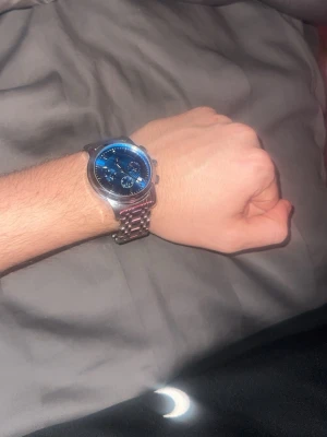 Silverfärgad klocka med blå urtavla - Säljer en stilren klocka med silverfärgat metallarmband och blå urtavla. Klockan har rund form och flera små visare på urtavlan. Perfekt för dig som vill ha en snygg accessoar till din outfit.