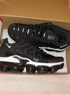 Nike Air Vapormax Plus svart/vit - Nike Air Vapormax Plus sneakers i svart med vita detaljer och synlig Air-sula. Ovandelen är i textil och syntet med vågigt mönster och små Swoosh-loggor. Skorna har snörning och en chunky, futuristisk design som sticker ut.