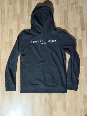 Svart hoodie från Tommy Hilfiger - Snygg svart hoodie från Tommy Hilfiger med klassisk logga och text på bröstet. Tröjan har en stor huva och en praktisk magficka. Detaljer i rött, vitt och blått på insidan av kragen ger en extra touch. Perfekt för en avslappnad och trendig stil.