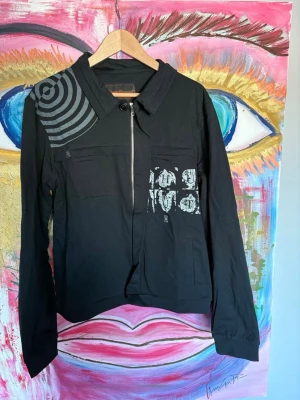 Number nine Jacka - Svart jacka med unikt grafiskt tryck i grått på axeln och bröstet. Jackan har en klassisk krage, dragkedja framtill och långa ärmar. Tillverkad i bomullsmaterial och har en modern, avslappnad passform. Perfekt för dig som gillar streetwear och vill sticka ut.