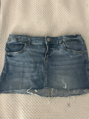 Blå jeanskjol från ONLY - Säljer en blå jeanskjol från ONLY med rå kant nertill och slitna detaljer. Kjolen har klassiska fem fickor, bälteshällor och knappstängning framtill. Perfekt för dig som gillar en avslappnad och trendig stil.