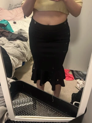 Svart volangkjol med tight passform - Säljer en svart kjol med volangkant nedtill och figurnära passform. Kjolen är midilång och har en enkel, stilren design som passar till många olika outfits. Perfekt för dig som gillar en klassisk men ändå trendig look.