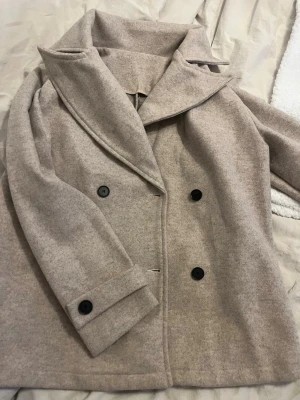 Beige dubbelknäppt ullkappa - Stilren beige kappa med dubbelknäppning och bred krage. Kappan har svarta knappar och är tillverkad i ett mjukt ullblandat material. Perfekt för dig som vill ha en klassisk och tidlös look med modern touch.