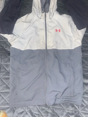 Under Armour vindjacka grå/vit - Snygg vindjacka från Under Armour i färgblockad design med grå nederdel, vita partier och svarta ärmar. Jackan har huva, dragkedja framtill, två sidofickor och Under Armour-logga i rött på bröstet. Perfekt för blåsiga dagar och sportiga outfits.
