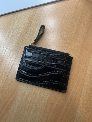 Svart plånbok med krokopräglat mönster - Snygg svart plånbok i konstläder med krokopräglat mönster och dragkedja upptill. Plånboken har ett handledsband i matchande material och flera kortfack på båda sidor. Perfekt för kort och kontanter när du vill resa lätt.