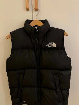 Svart dunväst från The North Face - Snygg svart dunväst från The North Face med hög krage och dragkedja framtill. Västen har två fickor med dragkedja och tydlig logga både fram och bak. Perfekt för lager-på-lager och riktigt varm tack vare dunfyllningen.