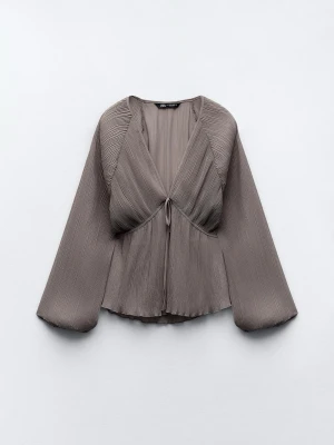 Plisserad taupe blus från Zara - Plisserad blus i taupe från Zara med v-ringning och knytning framtill. Blusen har vida långärmade ärmar och är tillverkad i ett lätt, glansigt material som ger en cool och modern look. Perfekt för dig som gillar trendiga detaljer och stilrena färger.