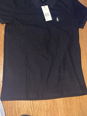 Svart piké från Polo Ralph Lauren - Klassisk svart pikétröja från Polo Ralph Lauren med vit broderad logga på bröstet. Tröjan har korta ärmar, krage och knappar vid halsen. Tillverkad i mjuk bomull som känns skön mot huden. Perfekt för en clean och stilren look.