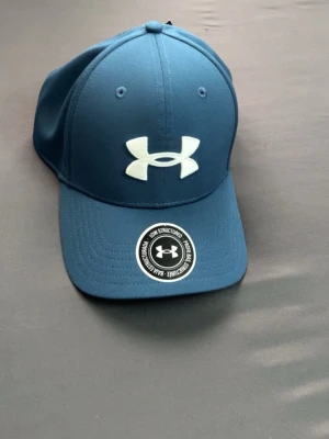 Blå keps från Under Armour - Snygg blå keps från Under Armour med böjd skärm och stort vitt logotryck framtill. Kepsen har klassisk sexpanelsdesign och justerbar baksida. Perfekt för en sportig och avslappnad stil.