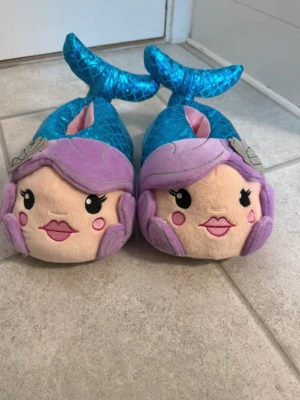 Blå glittriga sjöjungfru-tofflor - Supergulliga tofflor formade som sjöjungfrur med lila hår, ansikte framtill och blå glittrig fiskstjärt. Tillverkade i mjukt plyschmaterial med detaljer som snäckor och rosa kinder. Perfekta för dig som älskar fantasifulla accessoarer.