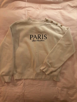 Beige sweatshirt med PARIS-tryck - Stilren beige sweatshirt med svart text 'PARIS Place Vendôme' på bröstet. Tröjan har rund halsringning och fyra dekorativa guldknappar på ena axeln. Mjukt material och ribbade muddar vid ärmslut och nederkant. Perfekt för en trendig och avslappnad look.
