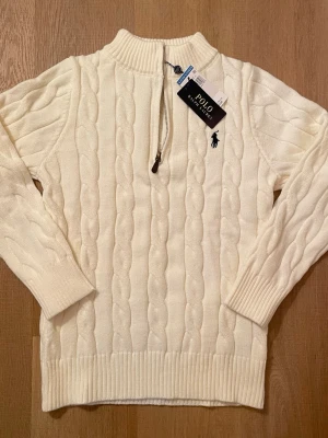 Vit kabelstickad tröja Polo Ralph Lauren - Kabelstickad vit tröja från Polo Ralph Lauren med halv dragkedja och hög krage. Tröjan har det klassiska broderade logotypen på bröstet och ribbade muddar vid ärmslut och nederkant. Perfekt för lager på lager och en clean look.