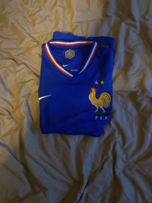 Blå Frankrike fotbollströja Nike - Snygg blå fotbollströja från Nike med Frankrikes landslagsmärke och två stjärnor på bröstet. Tröjan har V-ringning med röd, vit och blå kant samt korta ärmar. Tillverkad i lätt och ventilerande material, perfekt för träning eller match.