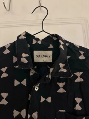 Svart skjorta Our Legacy med mönster - storlek 52/L - Svart långärmad skjorta från Our Legacy med vitt abstrakt mönster över hela plagget. Skjortan har klassisk krage, knäppning framtill och är tillverkad i mjuk bomull. Perfekt för dig som vill sticka ut lite extra.