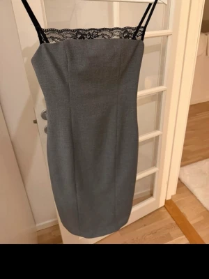 Grå axelbandsklänning med spets - Snygg grå kortklänning med svarta axelband och spetsdetalj upptill. Klänningen har en rak, figurnära passform och är ärmlös. Perfekt för dig som gillar stilrena och eleganta plagg med en twist.