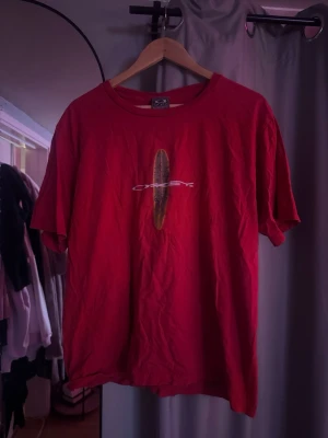 Röd Oakley t-shirt med tryck - Säljer en röd t-shirt från Oakley med ett coolt tryck av en surfbräda och Oakley-loggan på bröstet. T-shirten har rund hals och korta ärmar. Perfekt för dig som gillar streetwear och vill ha något unikt i garderoben.