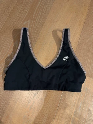 Svart sport-bh från Nike Air - Svart sport-bh från Nike Air med V-ringning och breda axelband. Kantbanden är beige med texten 'AIR' tryckt runtom. Bh:n har en stilren design och är tillverkad i ett stretchigt material som passar perfekt för träning.