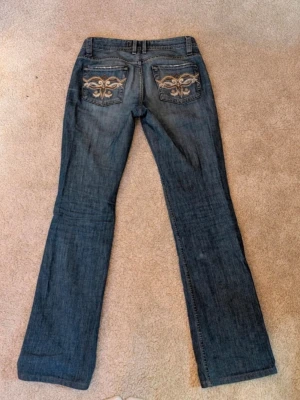 Blå bootcut jeans med brodyr - Snygga blå bootcut jeans med slitningar framtill och broderade detaljer på bakfickorna. Klassisk femficksmodell med låg midja och dragkedja. Perfekta till sneakers eller boots för en avslappnad look.