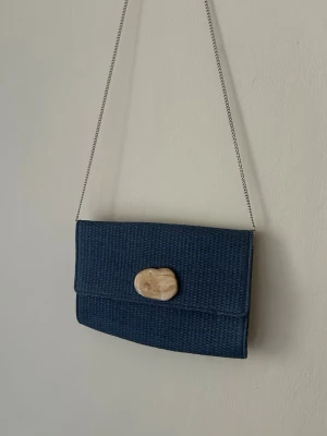 Mörkblå handväska med kedja - Stilren mörkblå kuvertväska med vävd struktur och silverfärgad kedjerem. Väskan har ett unikt, ljusbrunt dekorativt spänne framtill som ger en cool touch. Perfekt för dig som gillar minimalistisk design och vill sticka ut lite extra. Köpt i Frankrike. 