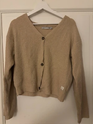 Beige ribbstickad kofta - Mysig beige ribbstickad kofta med v-ringning och tre knappar framtill. Koftan har en avslappnad passform, långa ärmar. Perfekt att slänga över en t-shirt eller linne.