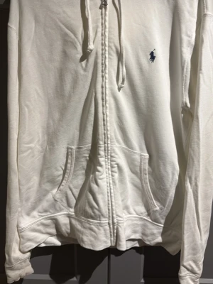 Vit hoodie från Polo Ralph Lauren - Säljer en stilren vit hoodie från Polo Ralph Lauren med dragkedja framtill och två fickor. Klassisk blå logga broderad på bröstet och justerbar huva med snören. Tillverkad i mjukt bomullsmaterial som känns skönt mot huden. Passar L   Som nyskick fast det är en otroligt liten fläck på ena armen (se bild 5)