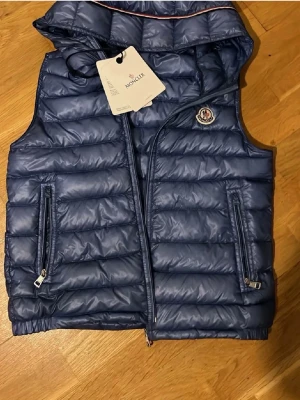 Blå väst från Moncler - Snygg blå dunväst från Moncler med huva och dragkedja framtill. Västen har två praktiska fickor med dragkedja och Moncler-logga på bröstet. Perfekt för lager-på-lager och ger en sportig vibe till din outfit.