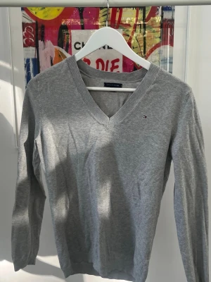 Grå V-ringad tröja Tommy Hilfiger - Klassisk grå V-ringad tröja från Tommy Hilfiger med liten broderad logga på bröstet. Tröjan har långa ärmar och ribbade muddar vid hals, ärmslut och nederkant. Perfekt för en stilren och enkel look. Storlek M i Junior dvs XS i Senior.