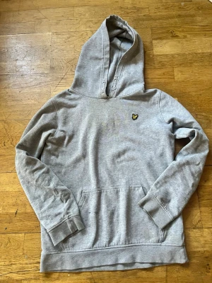 Grå hoodie från Lyle & Scott - Snygg grå hoodie från Lyle & Scott med klassisk gul logga på bröstet. Hoodien har en stor huva och en praktisk magficka. Tillverkad i mjukt bomullsmaterial och har ribbade muddar vid ärmslut och nederkant. Perfekt för en avslappnad stil.