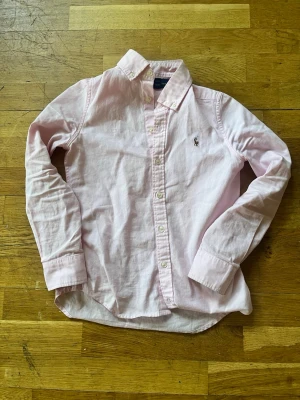 Ljusrosa skjorta från Ralph Lauren - Klassisk ljusrosa skjorta från Ralph Lauren med broderad logga på bröstet. Skjortan har knappar framtill, lång ärm och en tidlös krage. Perfekt för en stilren look och tillverkad i mjuk bomull.