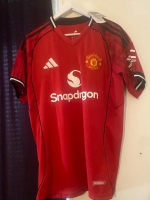 Manchester United matchtröja röd Adidas - Röd Manchester United matchtröja från Adidas med klubbmärke på bröstet, vita loggor och svarta detaljer. Tröjan har V-ringning, svarta ränder på axlarna och är tillverkad i lätt funktionsmaterial (AEROREADY). Perfekt för fotboll eller träning.