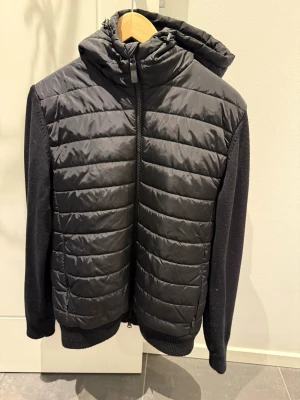 Svart cardigan från J.Lindeberg - Snygg svart pufferjacka från J.Lindeberg med quiltad framsida och huva. Ärmarna är stickade i mjukt material och jackan har dragkedja framtill. Perfekt för kalla dagar och ger en sportig look.