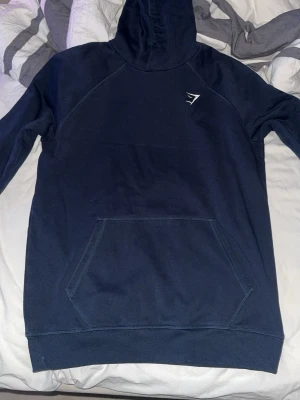Mörkblå hoodie från Gymshark - Mörkblå hoodie från Gymshark med klassisk huva och stor magficka. Diskret Gymshark-logga på bröstet. Perfekt för chill eller träning. Mjuk bomullskänsla och snygg passform.